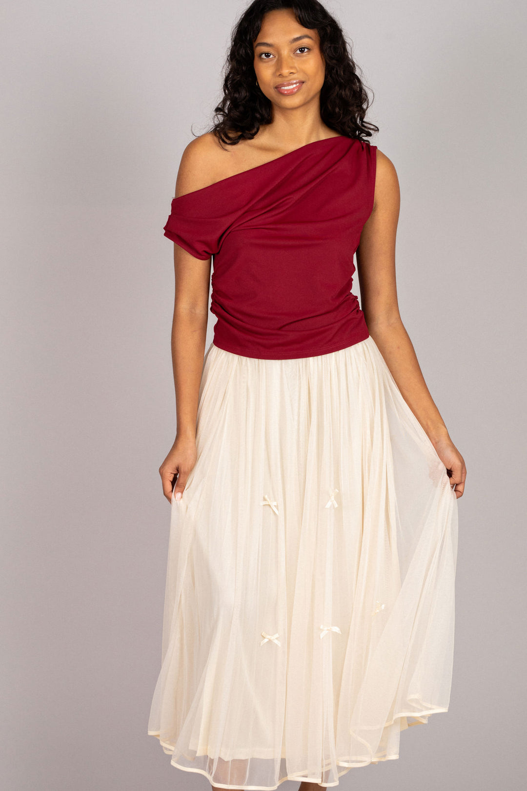 A sleeveless red top with an asymmetrical neckline and a cream pleated midi skirt, worn by a smiling woman with curly dark hair.