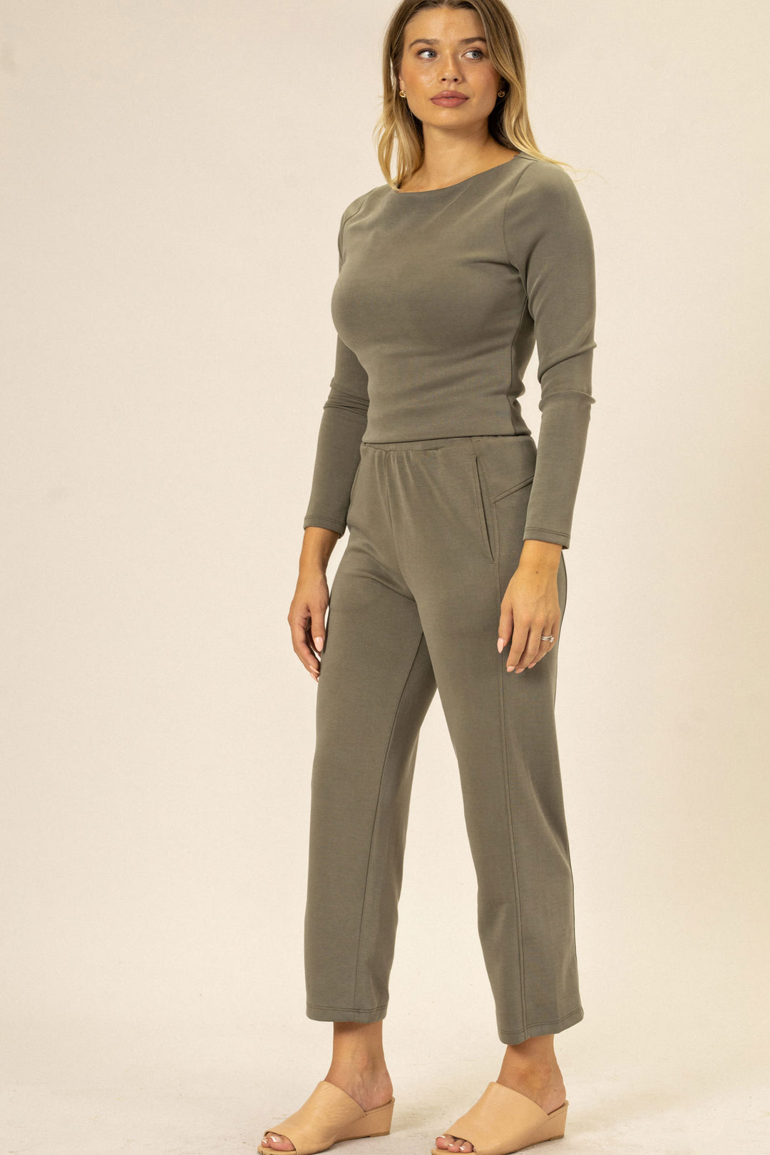 A woman wearing a gray long-sleeved jersey top and straight-leg pants, with pockets, standing against a beige background.
