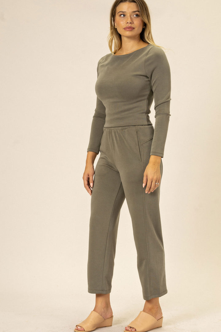 A woman wearing a gray long-sleeved jersey top and straight-leg pants, with pockets, standing against a beige background.