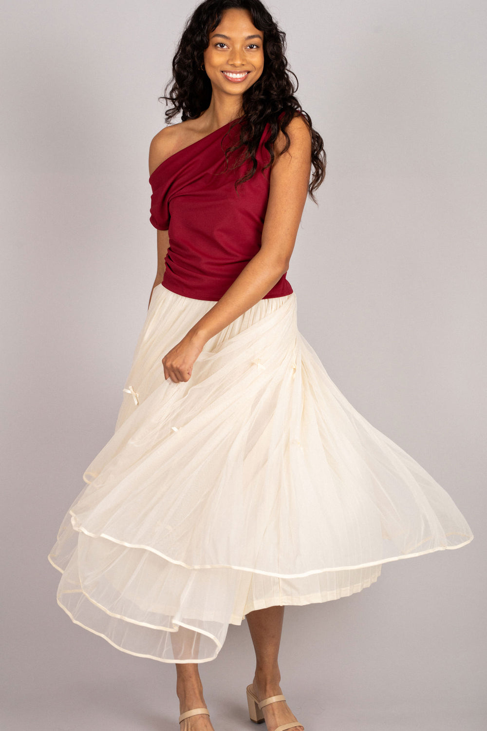 A model wearing a red asymmetrical ruched top and a long white pleated skirt, smiling confidently.