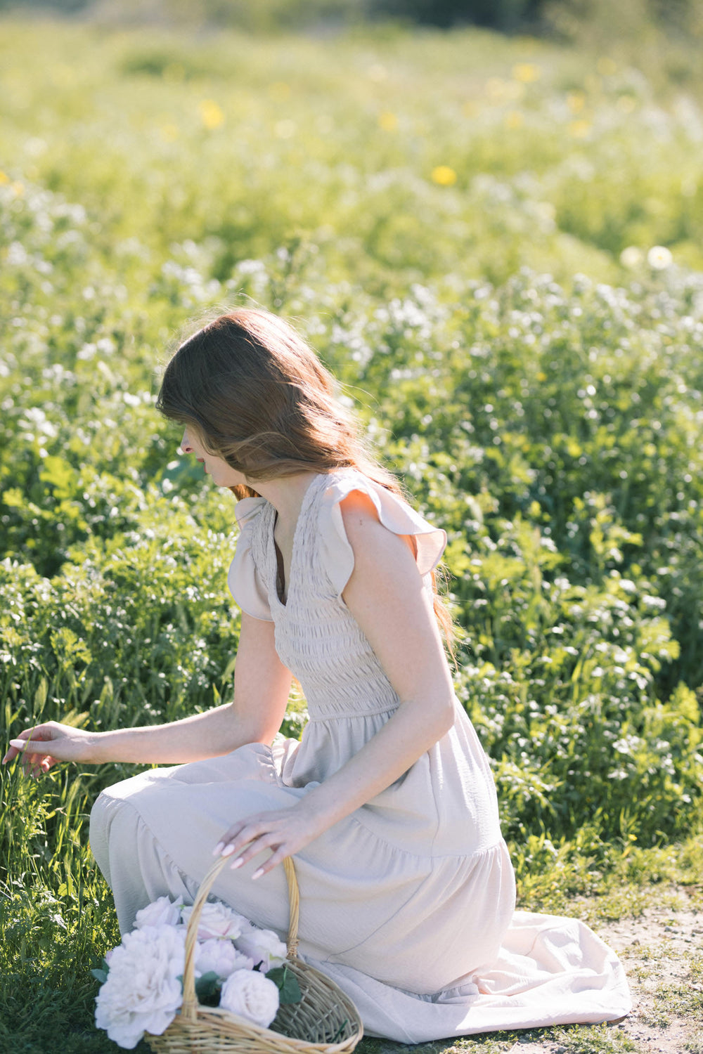 A young woman wearing a white smocked v-neck dress sits in a field of yellow and white wildflowers, holding a basket of flowers.