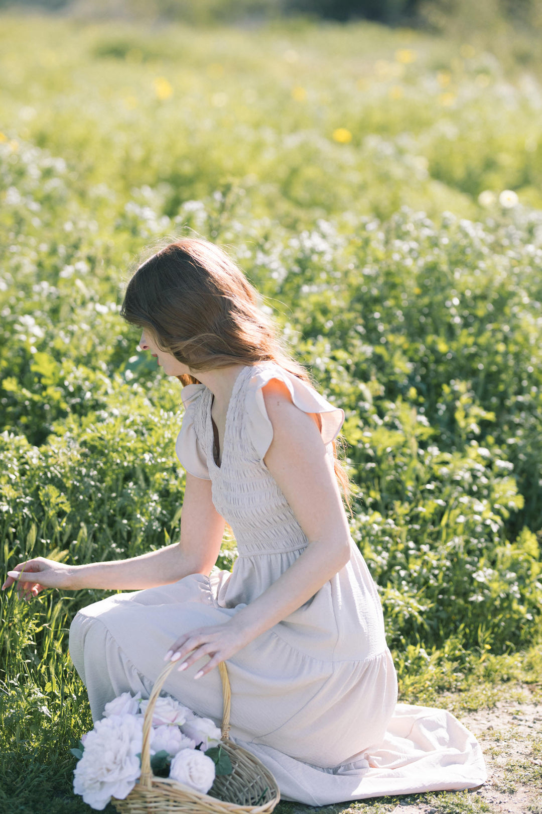 A young woman wearing a white smocked v-neck dress sits in a field of yellow and white wildflowers, holding a basket of flowers.