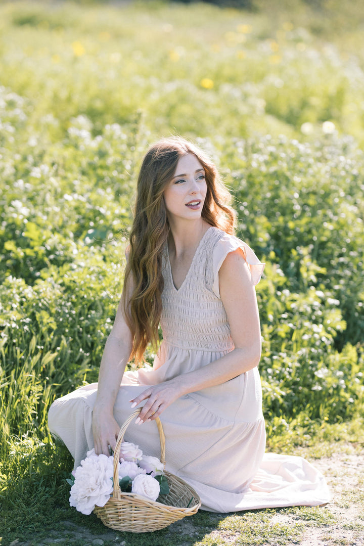 A woman in a long, light gray dress stands in a field of white flowers, holding a wicker basket filled with white peonies.