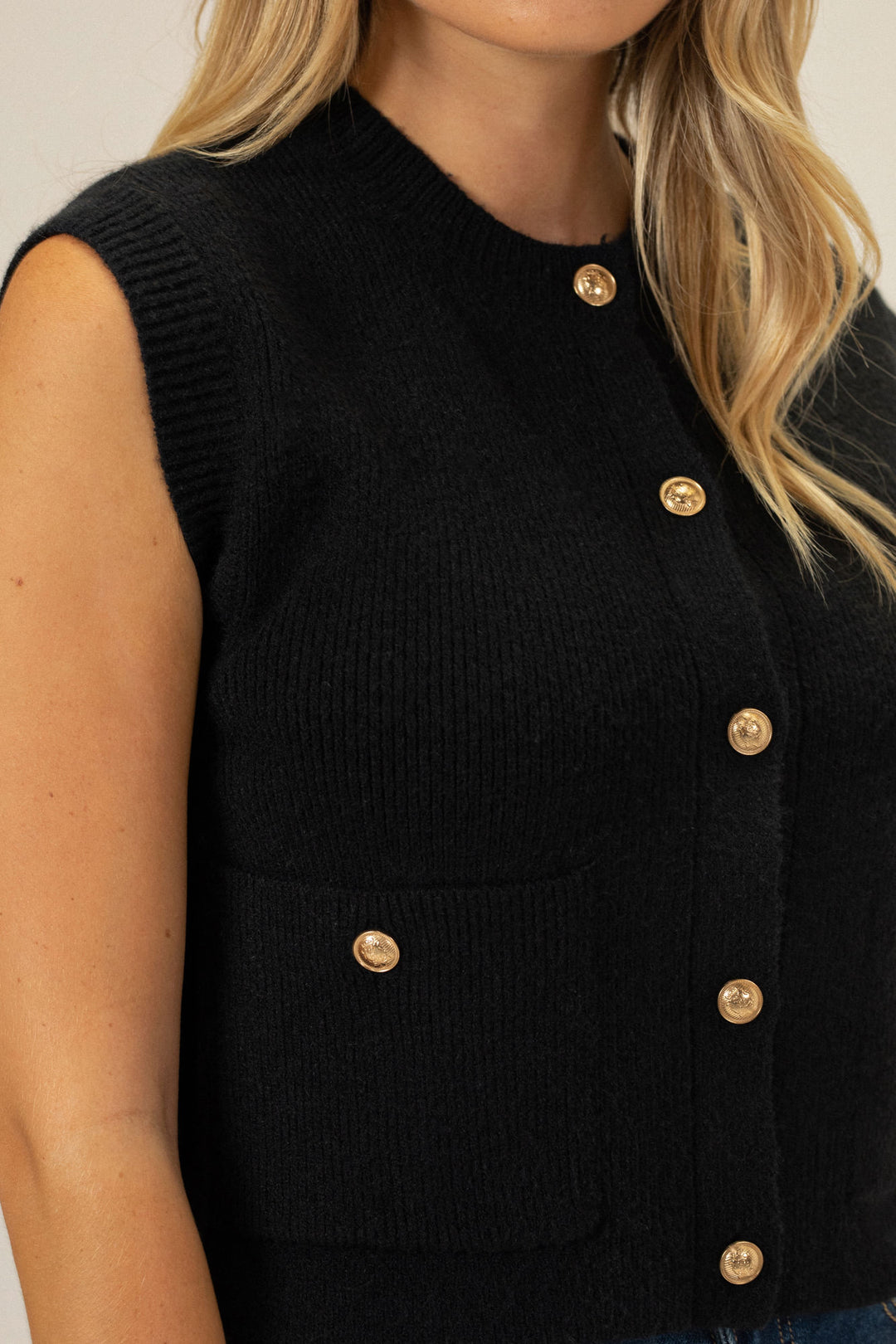 A black knit vest with a high collar, gold buttons, and ribbed detailing, worn by a model with long blond hair.