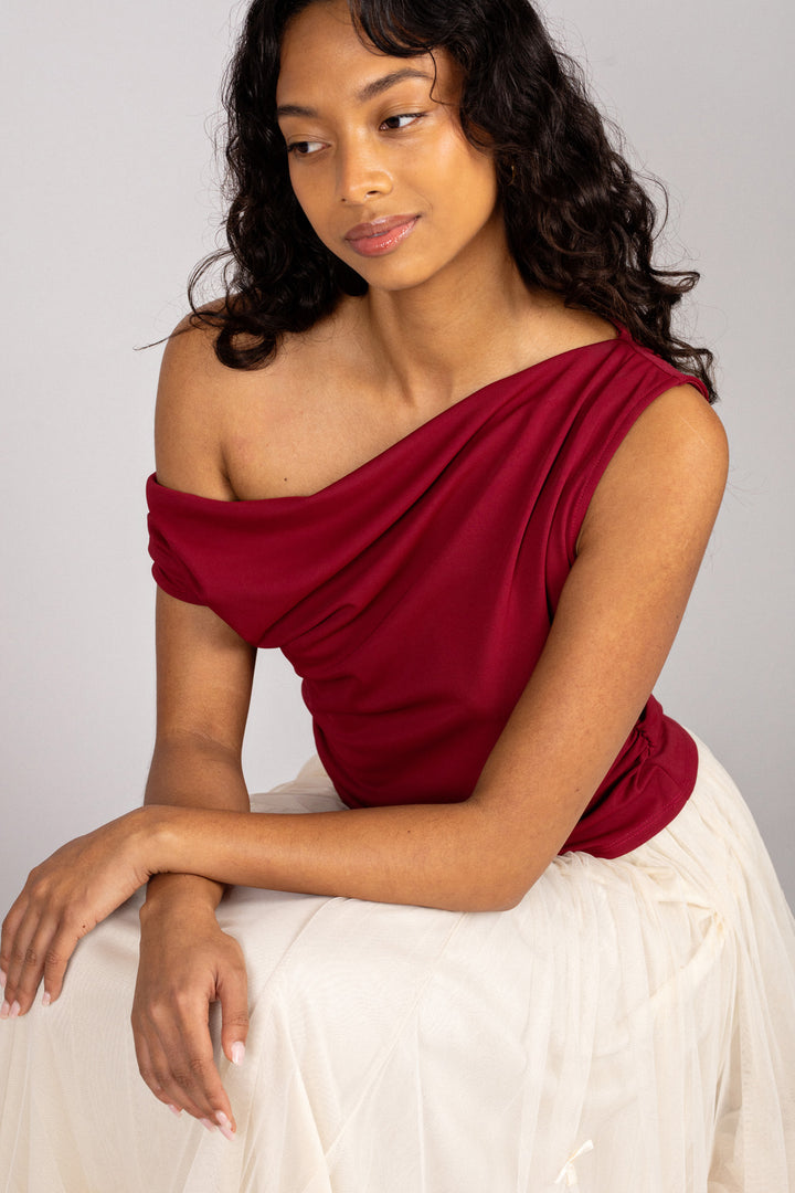 A woman wearing a red, asymmetrical ruched top with a draped neckline, paired with a white skirt, showcasing the style and fit of the Lexie top.
