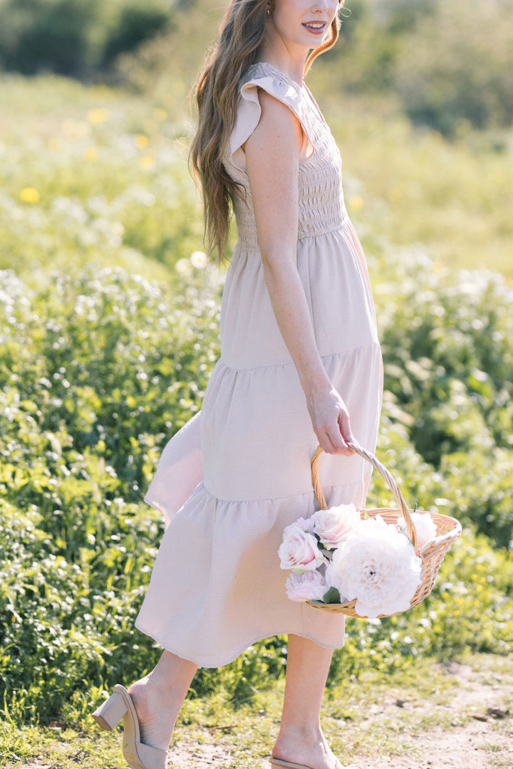 A woman in a light gray smocked midi dress holding a basket of pink flowers, standing in a field of yellow flowers.