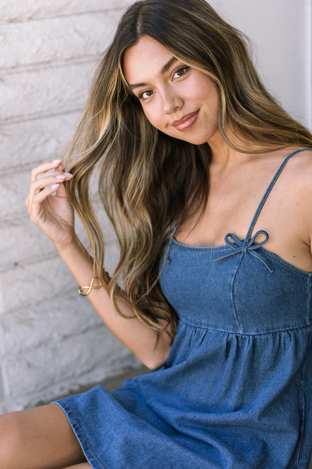 Young woman with long wavy brown hair, wearing a blue denim mini dress with a bow detail at the chest, standing against a white wall.