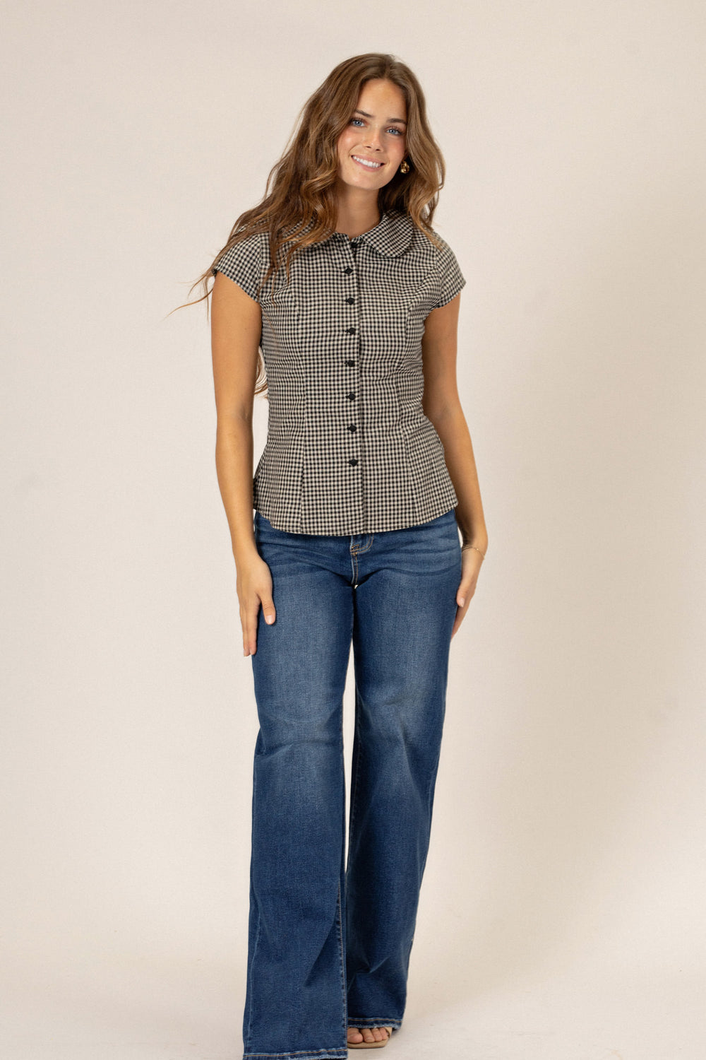 A young woman in a fitted black-and-white gingham button-up shirt, paired with blue jeans, smiling at the camera.
