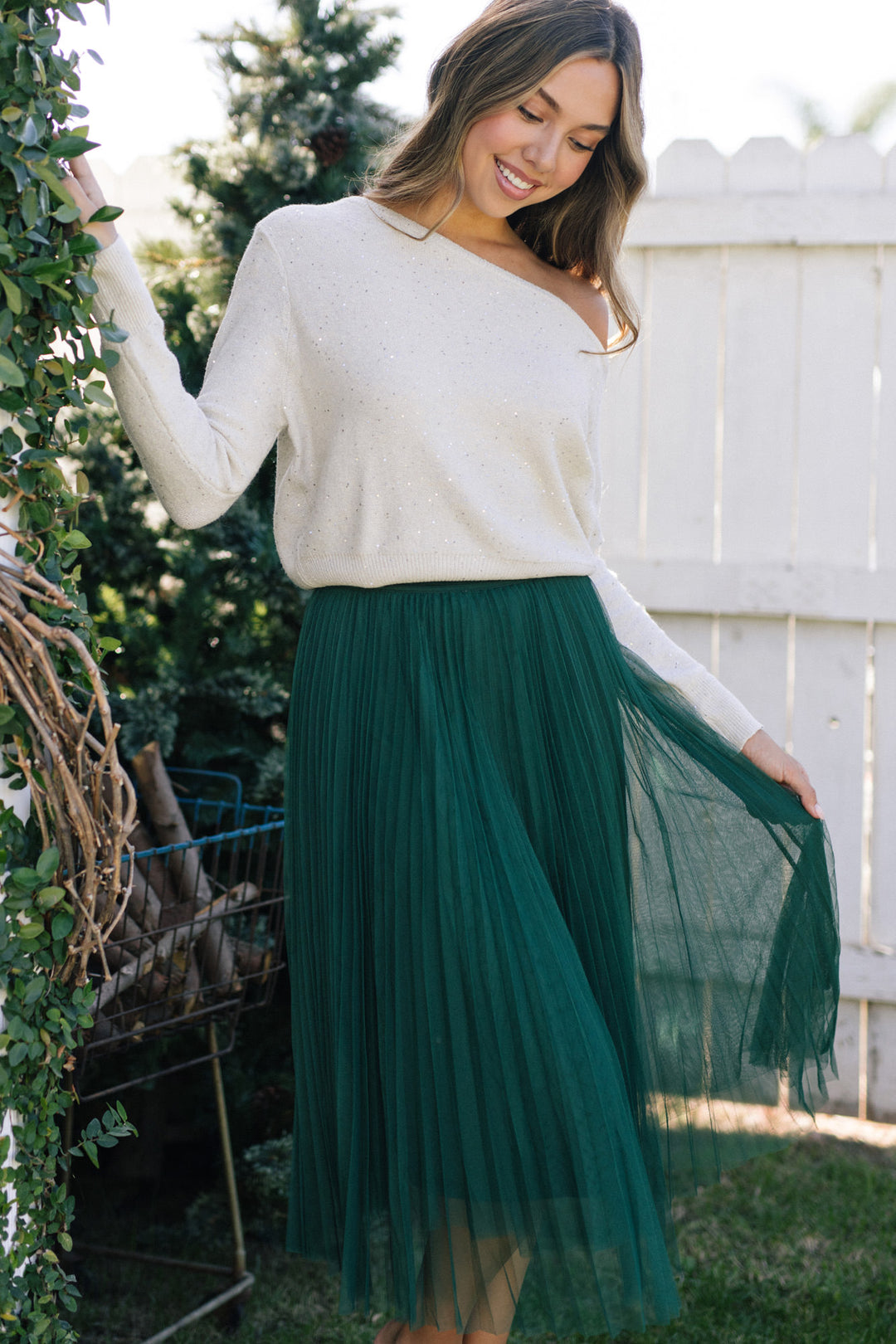 A smiling woman wearing a white, speckled knit sweater and a flowing, emerald green tulle skirt, posing against a backdrop of greenery.