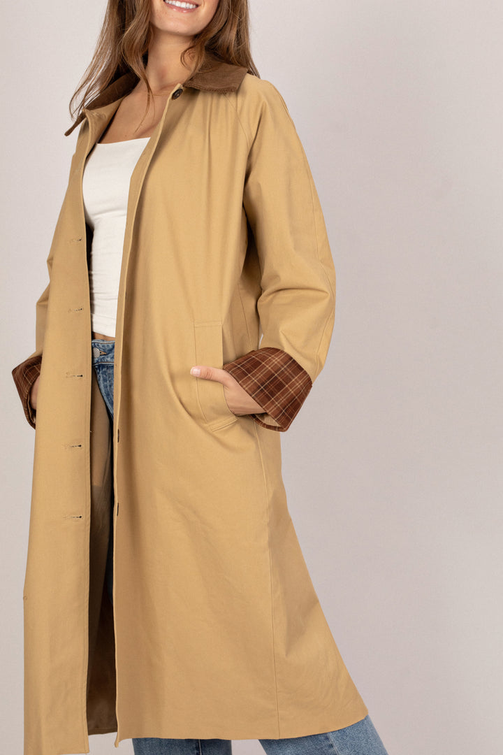 The image shows a woman wearing a tan, knee-length coat with a corduroy collar and cuffs. The coat has a relaxed, casual style with functional pockets.