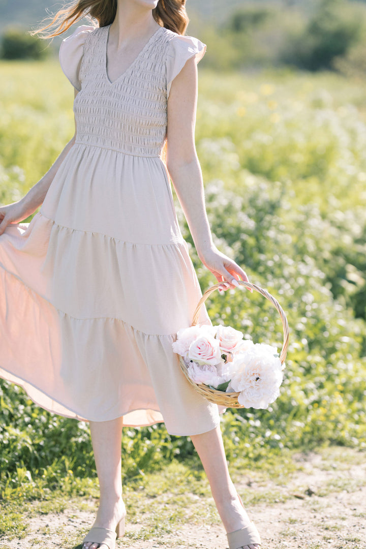 A smocked, V-neck dress in a soft, neutral tone with a flowing, tiered skirt, carried in a wicker basket filled with lush white flowers.