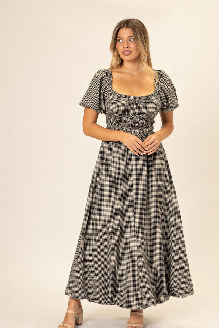 A model wearing a black and white gingham dress with a fitted bodice, puffed sleeves, and a full, flared skirt.