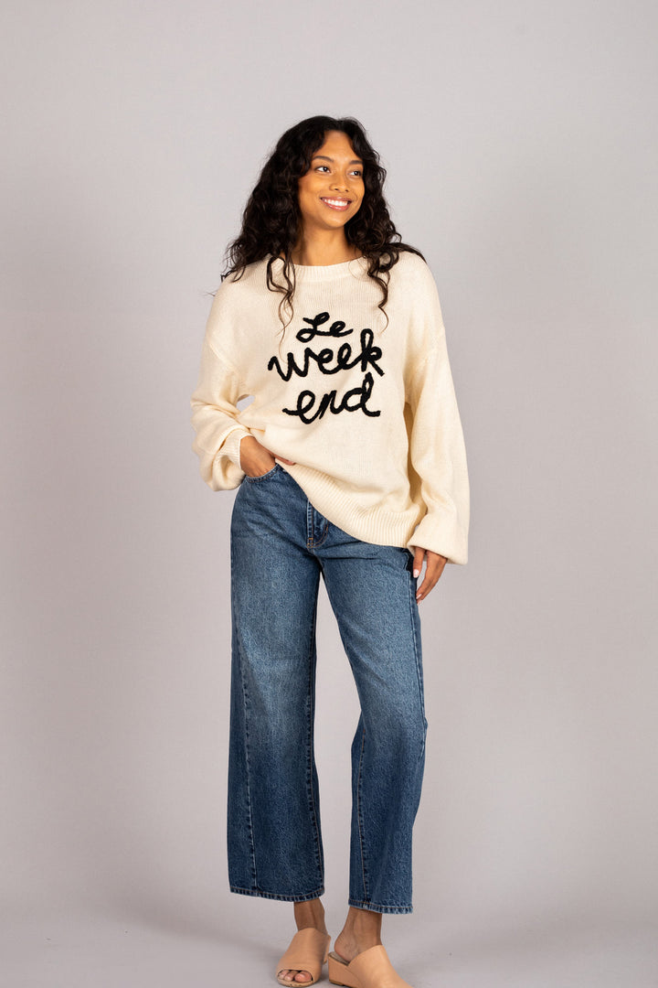 A young woman wearing a cream-colored knit sweater with the text "Le week end" embroidered on the front, paired with high-waisted denim jeans and beige sandals, standing against a light gray background.