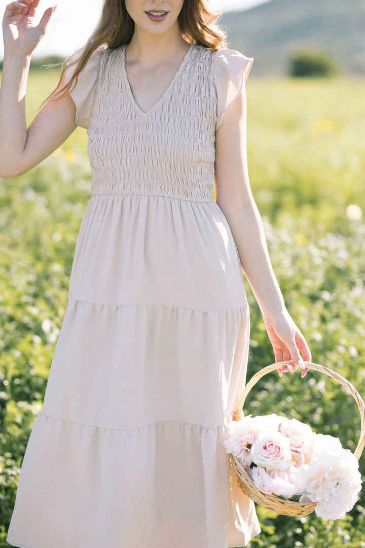 A smocked, V-neck dress in a light neutral color, held by a woman in a field of flowers, with a woven basket of flowers in her hand.