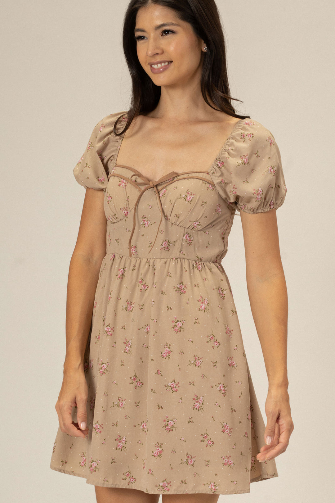 The image shows a young woman wearing a beige floral-patterned mini dress with puff sleeves and a ruffled bodice, creating a romantic, feminine look.