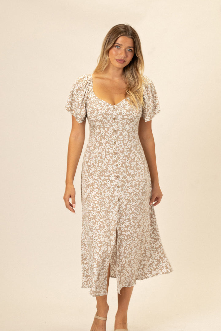 Model wearing a long, fitted floral dress with a v-neck and puffed sleeves in a light, airy color palette.
