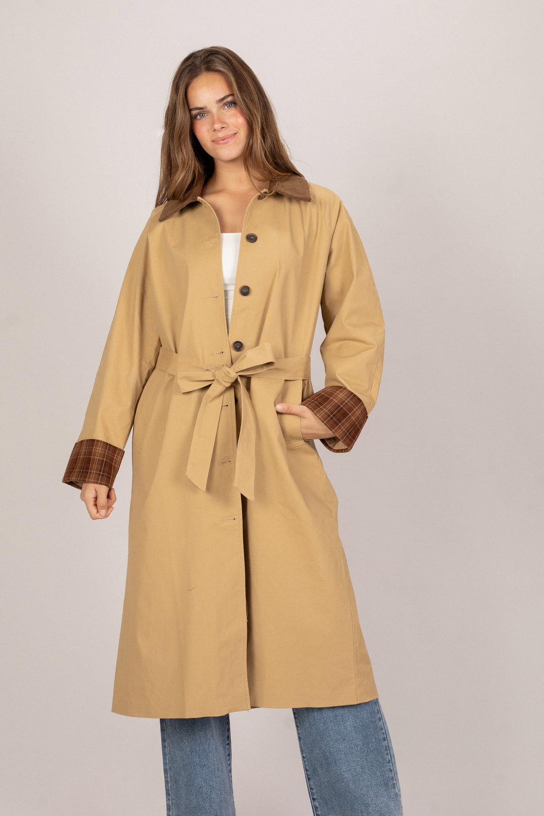 A beige trench coat with a corduroy collar, a belted waist, and plaid cuffs, modeled by a woman with long brown hair.