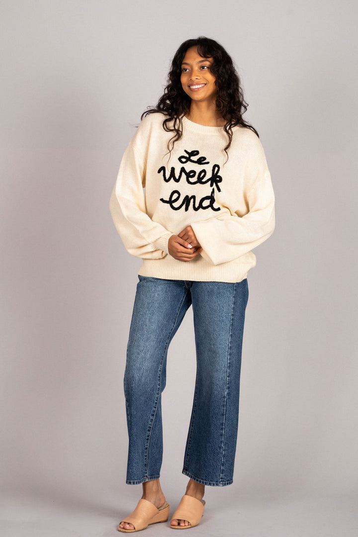 Cream-colored sweater with embroidered "weekend" text, worn by a smiling woman with curly hair and jeans.