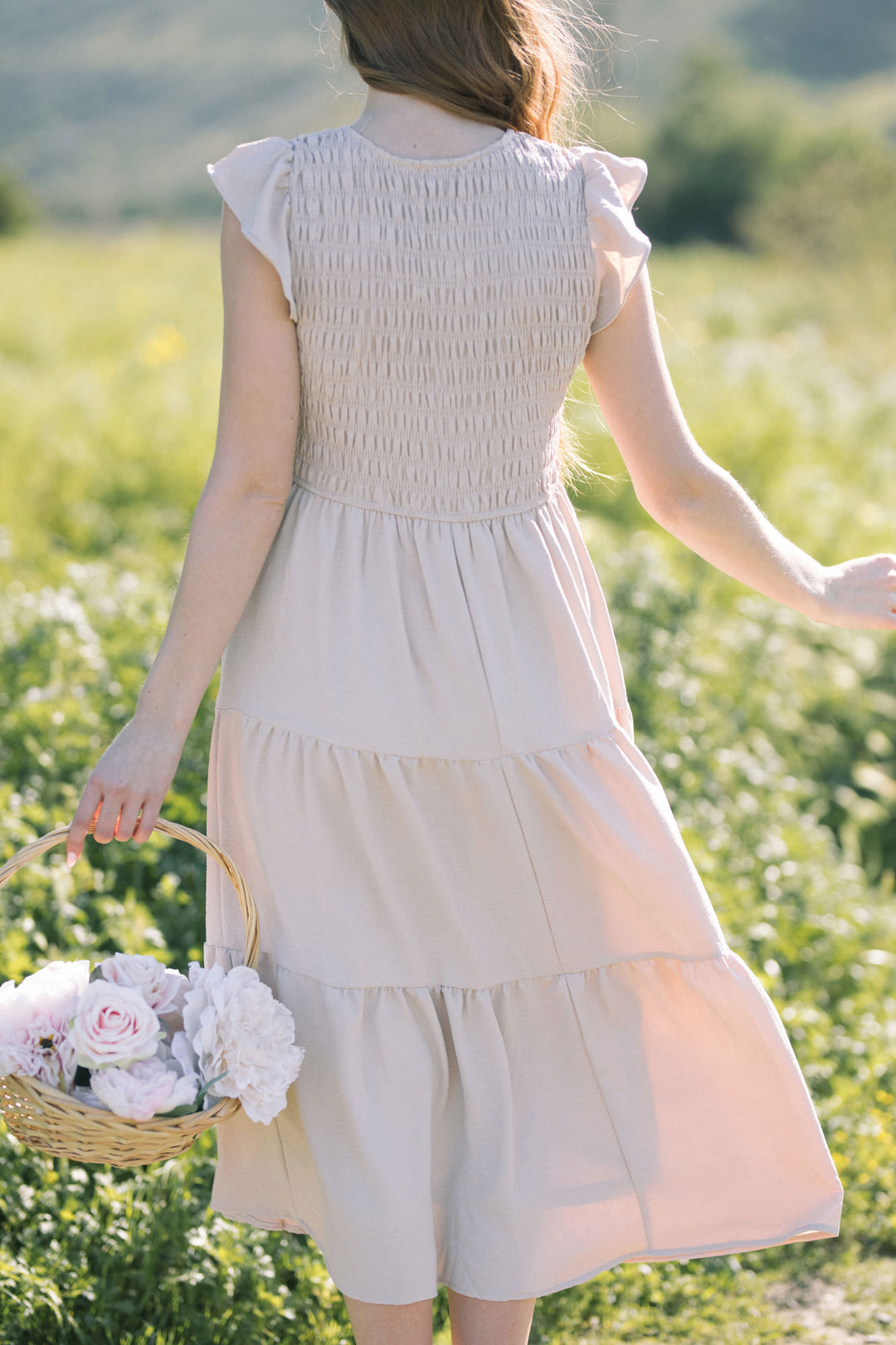 A white, smocked-bodice dress with tiered, flowy skirt and ruffled sleeves, held in a woman's hand against a blooming field.