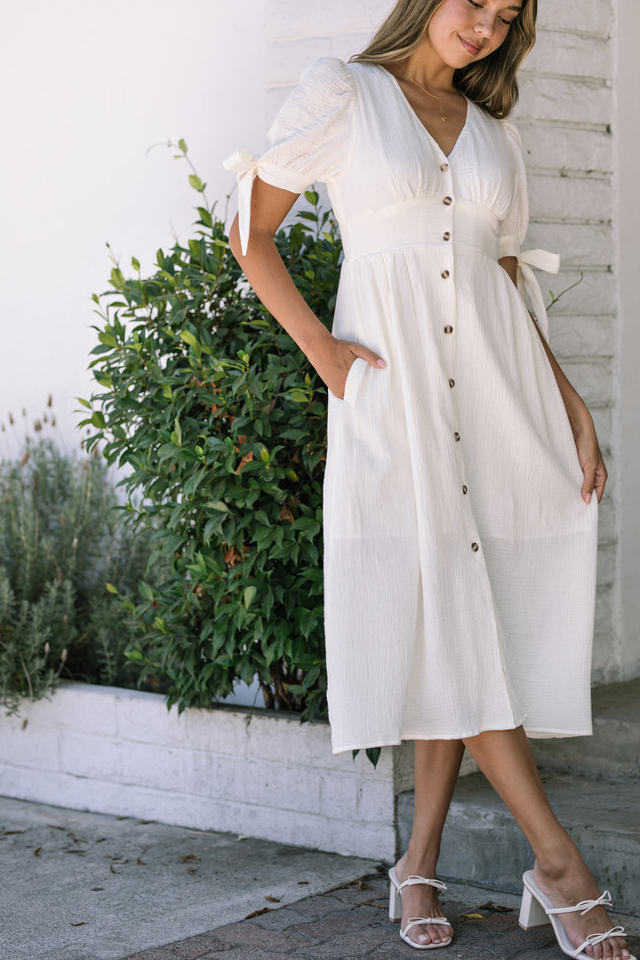 A white, midi-length dress with puffed sleeves, a v-neck, and a row of buttons down the front, worn by a woman with long brown hair standing in front of greenery.