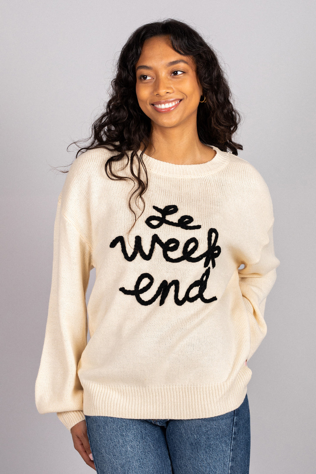 Cream-colored knit sweater with embroidered text "Le Weekend End" worn by a smiling, curly-haired woman.