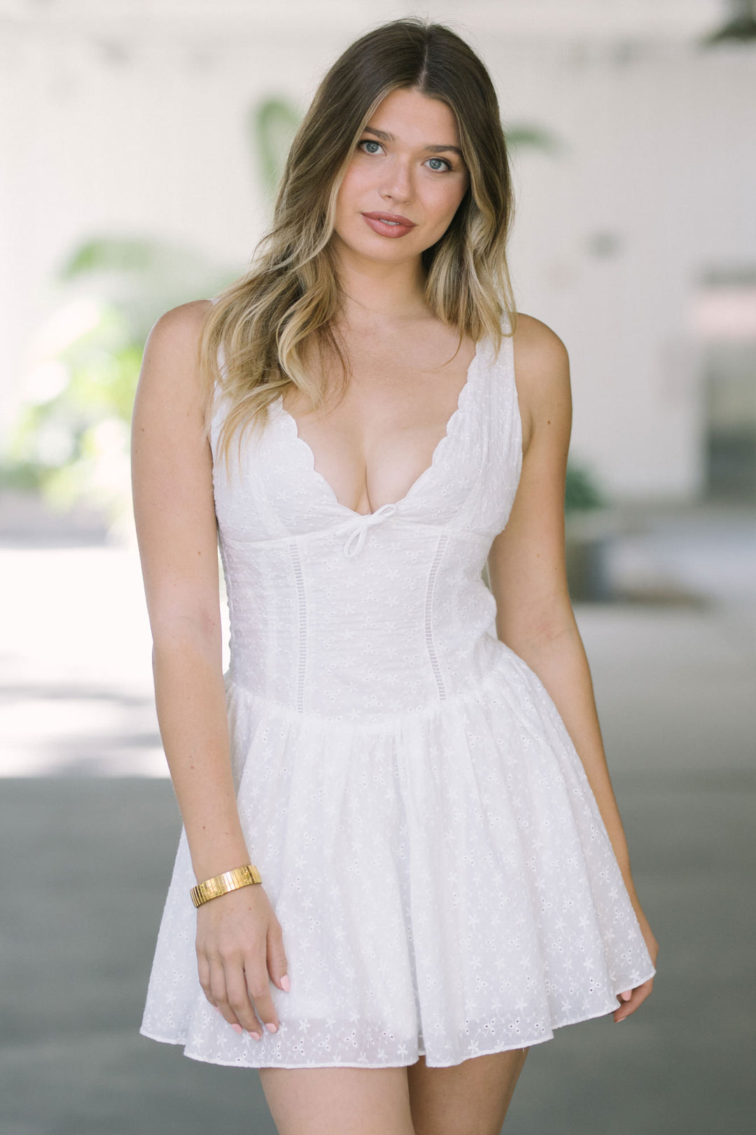 A white sleeveless mini dress with scalloped eyelet lace detailing, featuring a plunging neckline and a flared, flowy silhouette.