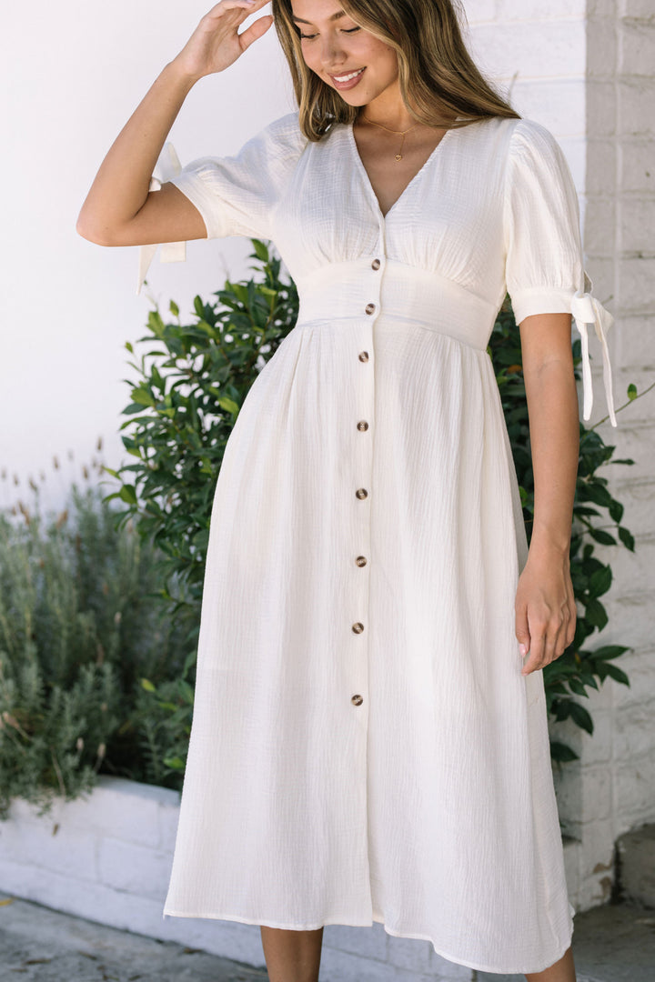 A white, midi-length dress with a V-neck, puffed sleeves, and a button-down front, creating a feminine, flowy silhouette.