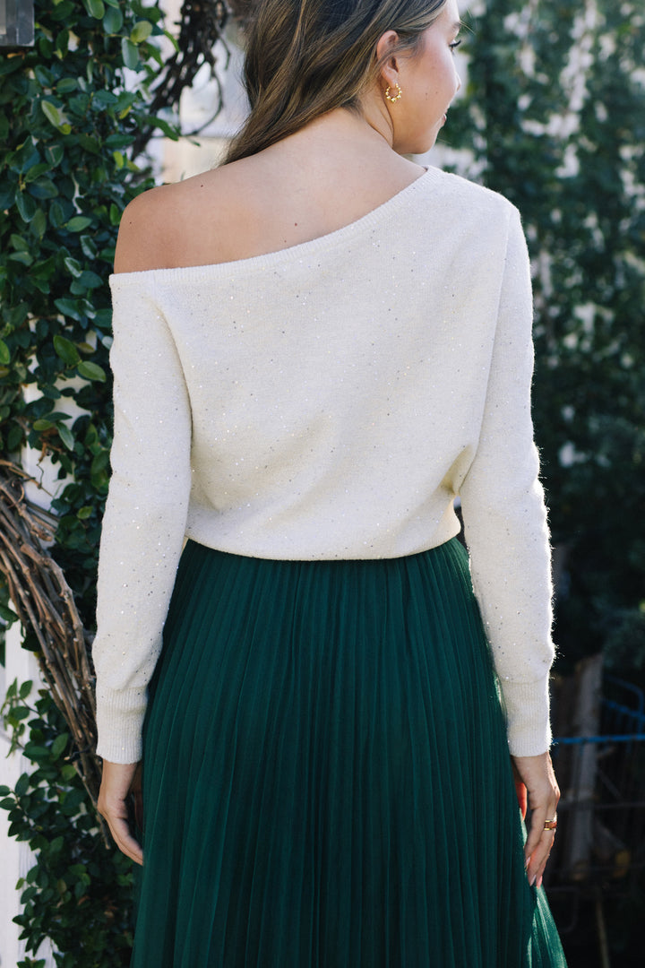 A woman wearing a white sequin-embellished knit sweater with an off-the-shoulder neckline, paired with a long, pleated emerald green skirt, set against a lush outdoor backdrop.