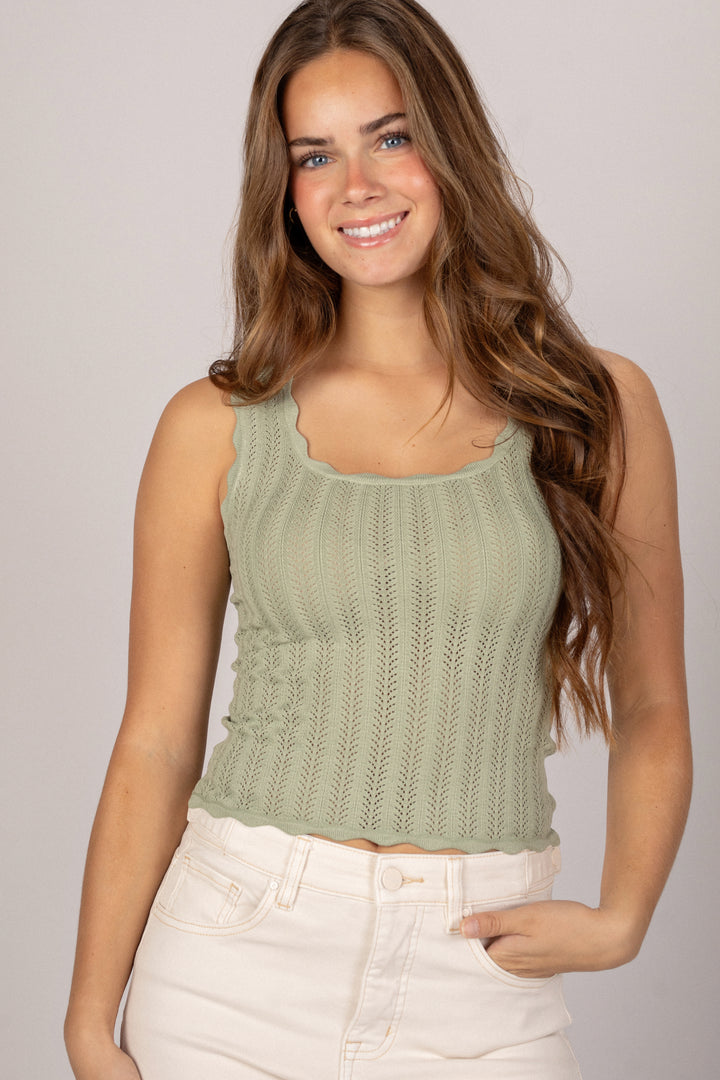 A woman with long, wavy brown hair wearing a scalloped, pointelle knit tank top in a light olive green color.