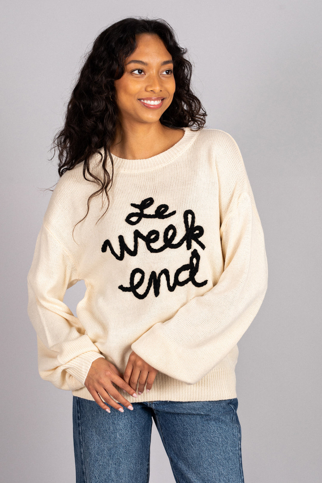 A woman wearing a cream-colored, knit sweater with "Le week end" embroidered in black text, paired with blue jeans.