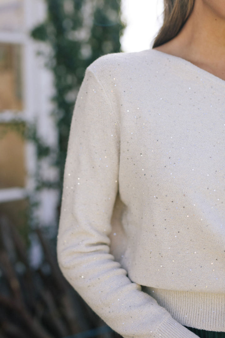 A close-up view of the back of a cream-colored knitted sweater with delicate sequin details scattered across the fabric, creating a subtle sparkling effect.