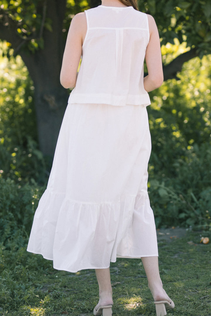 A white, sleeveless midi dress with a ruffle hem and a tie detail at the front, set against a lush green backdrop.