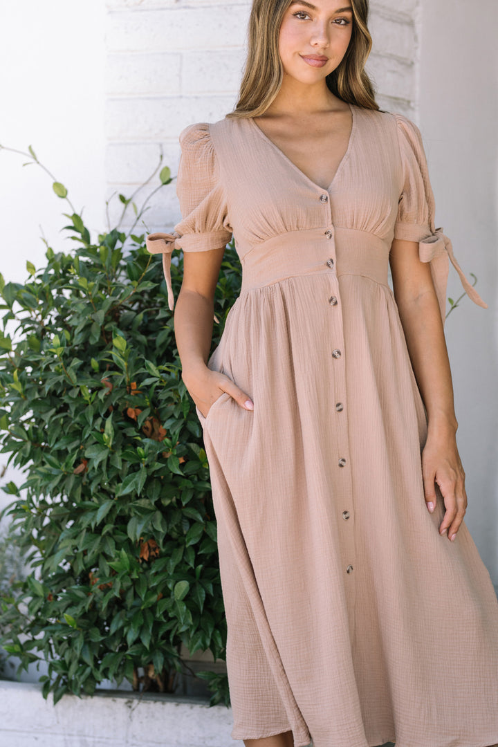 A beige, pleated gauze dress with puffed sleeves, waist seam, and button front closure, modeled by a woman with long brown hair.