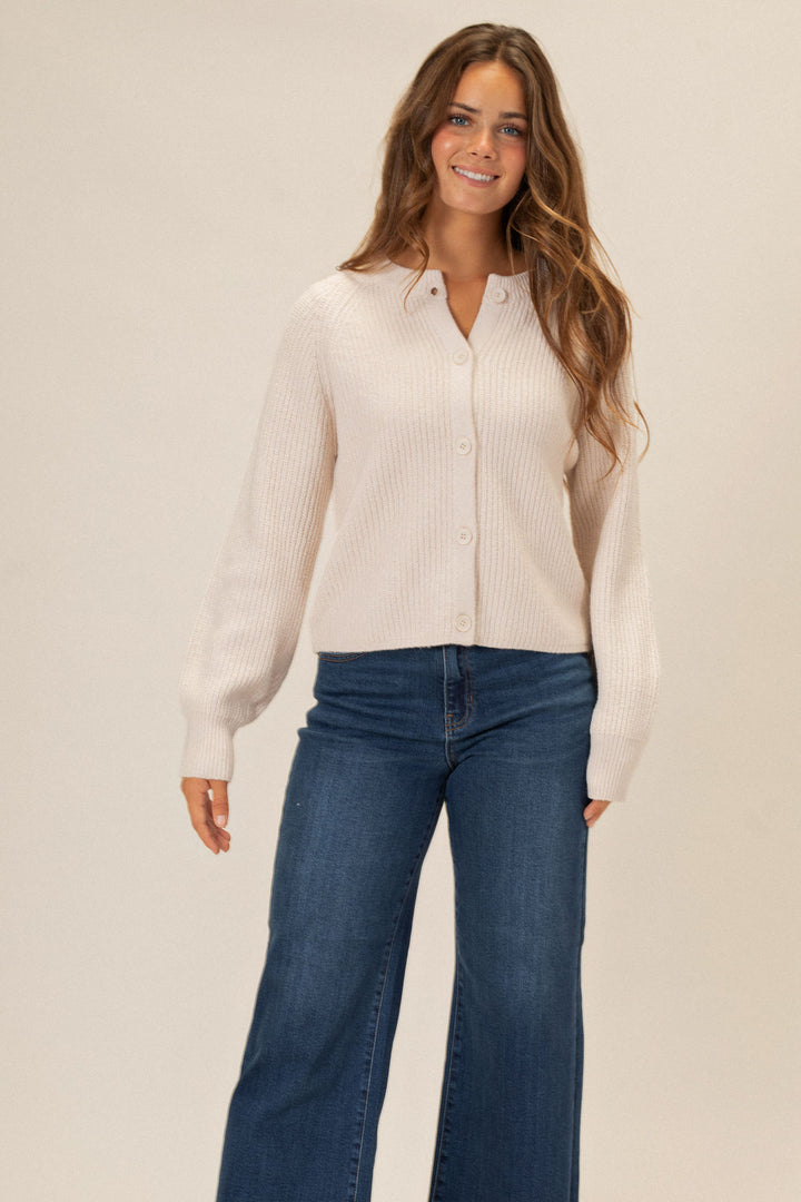 A young woman in a cozy, ribbed white knit cardigan with button front, posing in jeans against a light background.