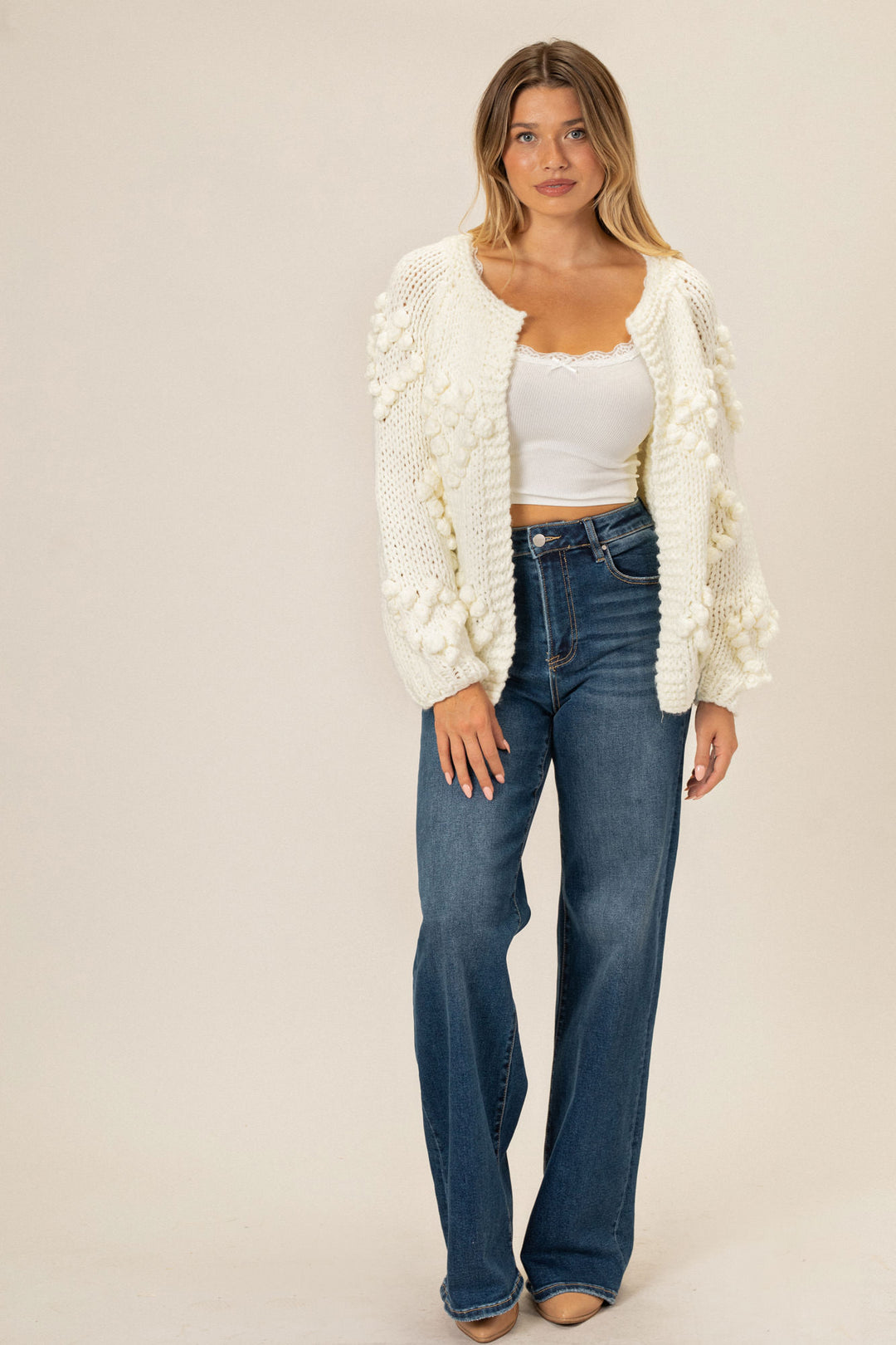 A woman wearing a white, knit cardigan with pom-pom details, paired with high-waisted denim jeans.