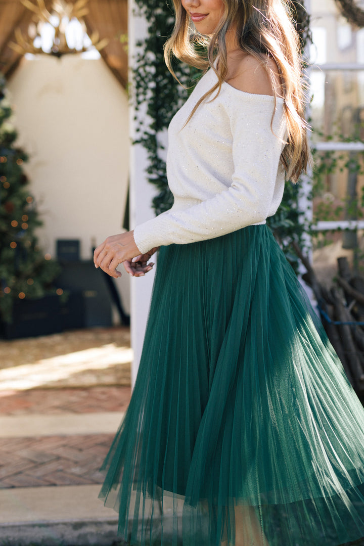 A young woman wearing a white sequin-embellished knit sweater with an off-the-shoulder neckline and a flowing, pleated emerald green midi skirt.