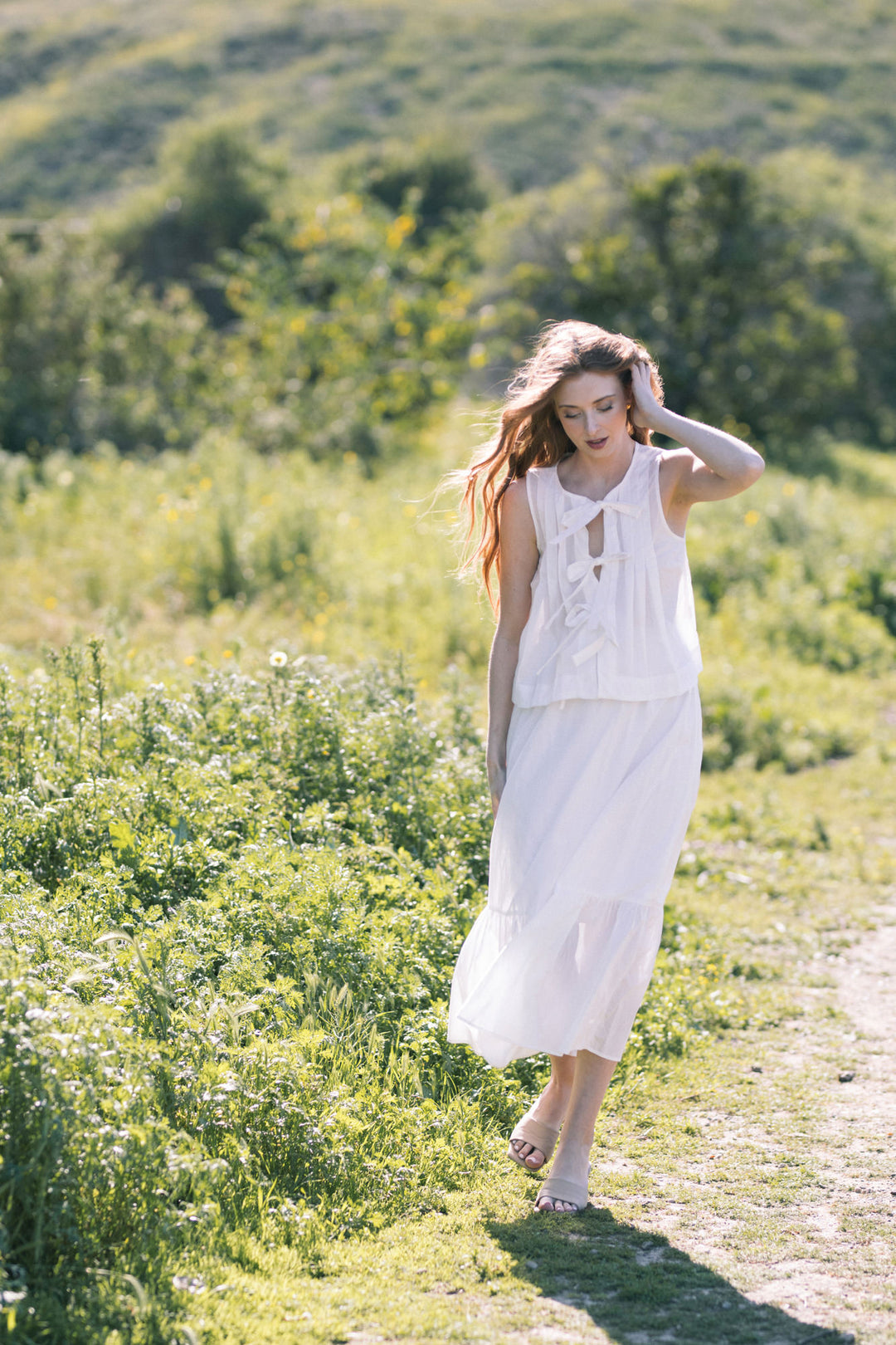 A woman in a long white dress walking through a lush green field with yellow flowers, her hands in her hair as the wind blows through her wavy hair.