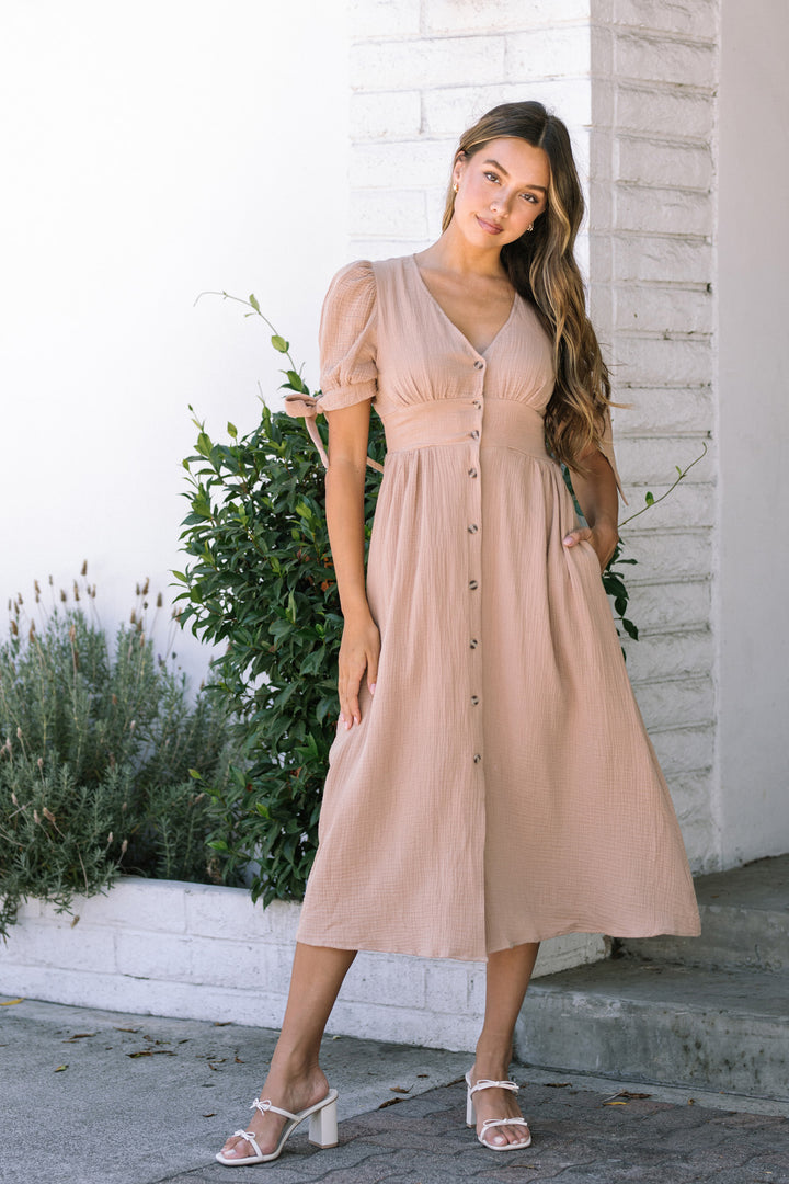A light peach-colored, puff-sleeved dress with a v-neckline, button front, and pleated midi skirt, worn by a tall, slim model against a white brick wall.