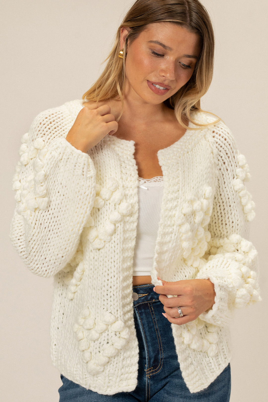 Knitted white cardigan with pom-pom details, worn by model with blonde hair and jeans.