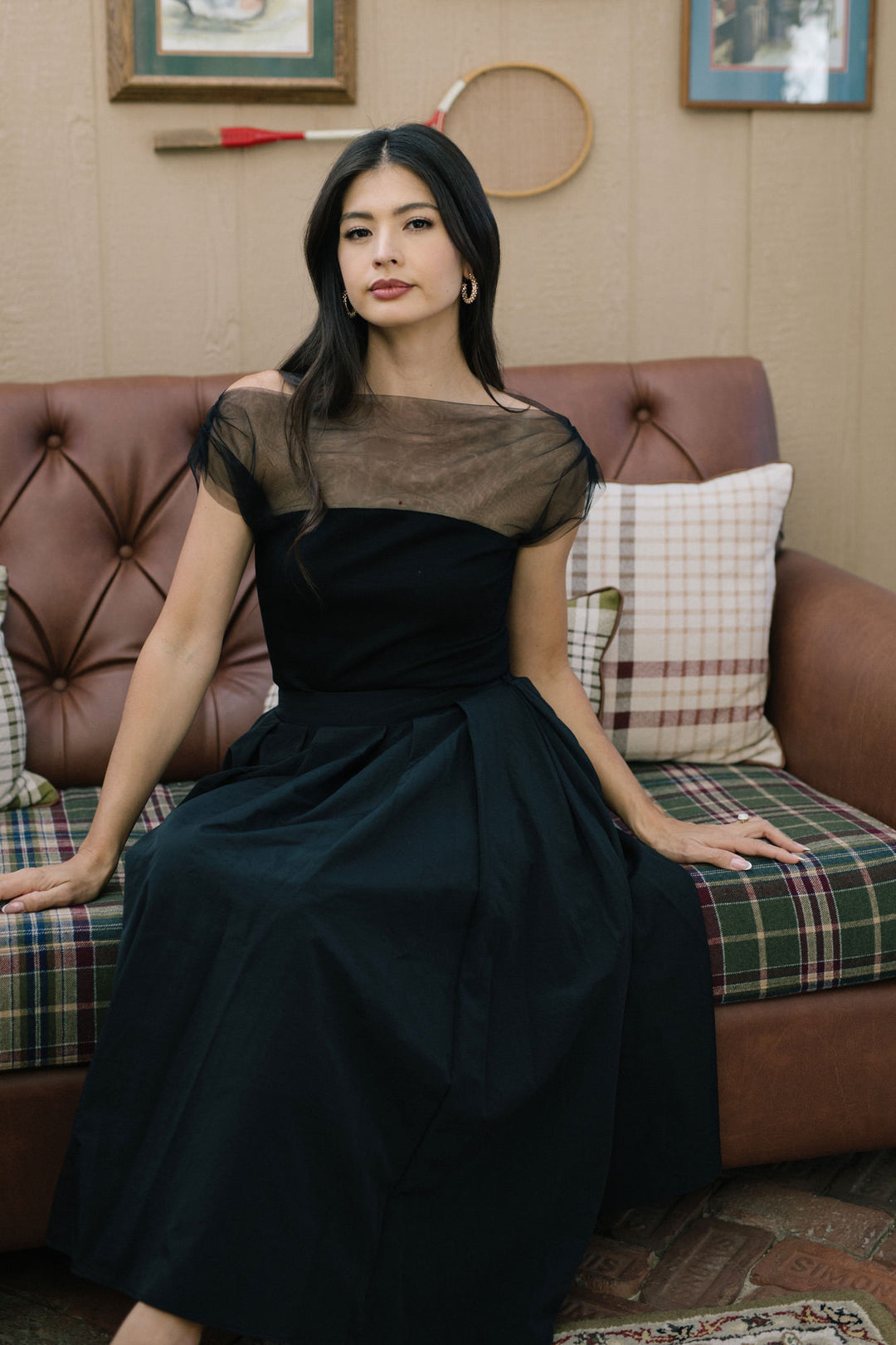 A young Asian woman with long, dark hair is seated on a patterned couch, wearing a black dress with a sheer mesh overlay on the upper bodice.