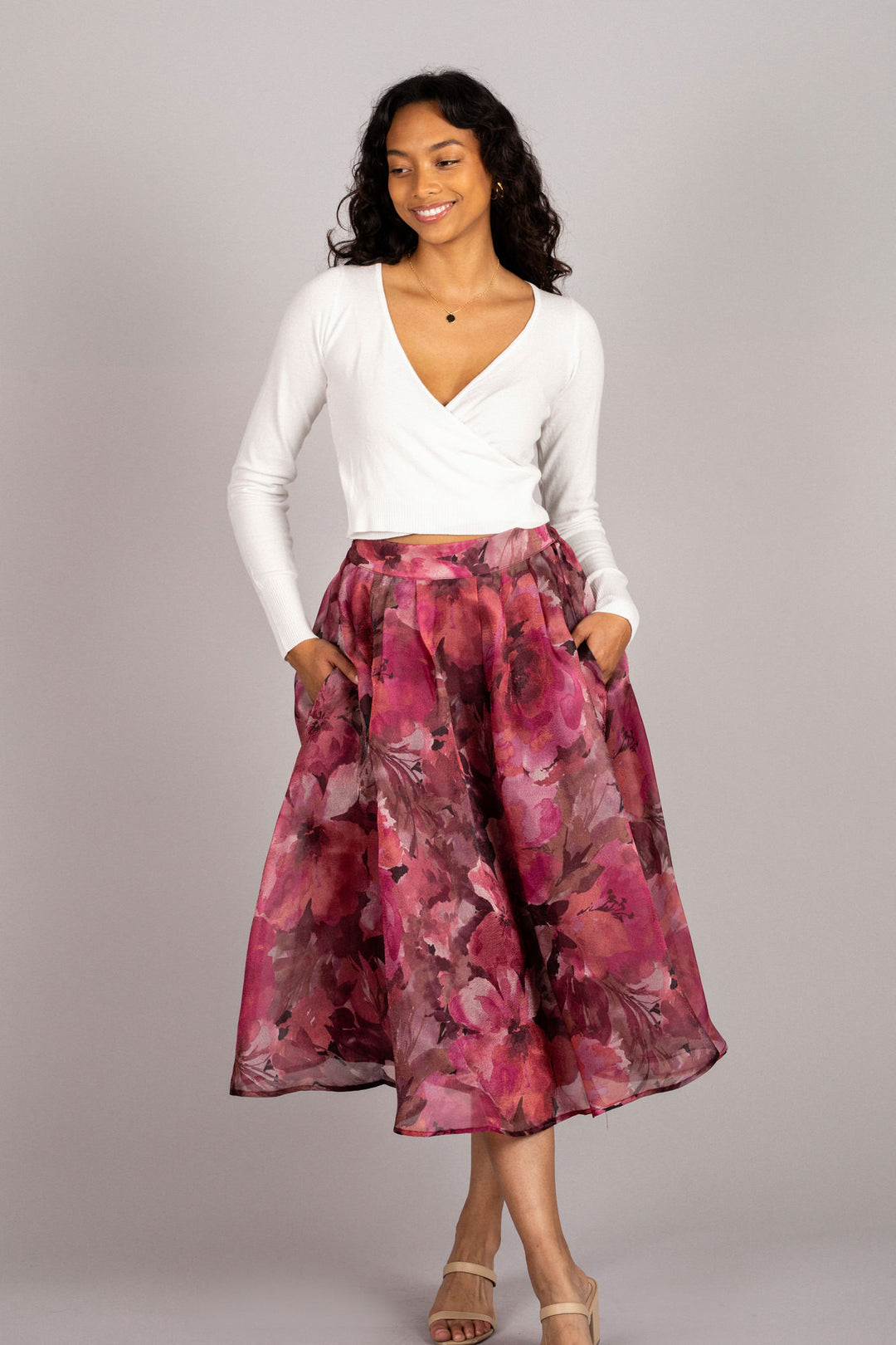 A woman wearing a white cropped top and a vibrant, floral midi skirt in shades of pink and red, posing against a gray background.