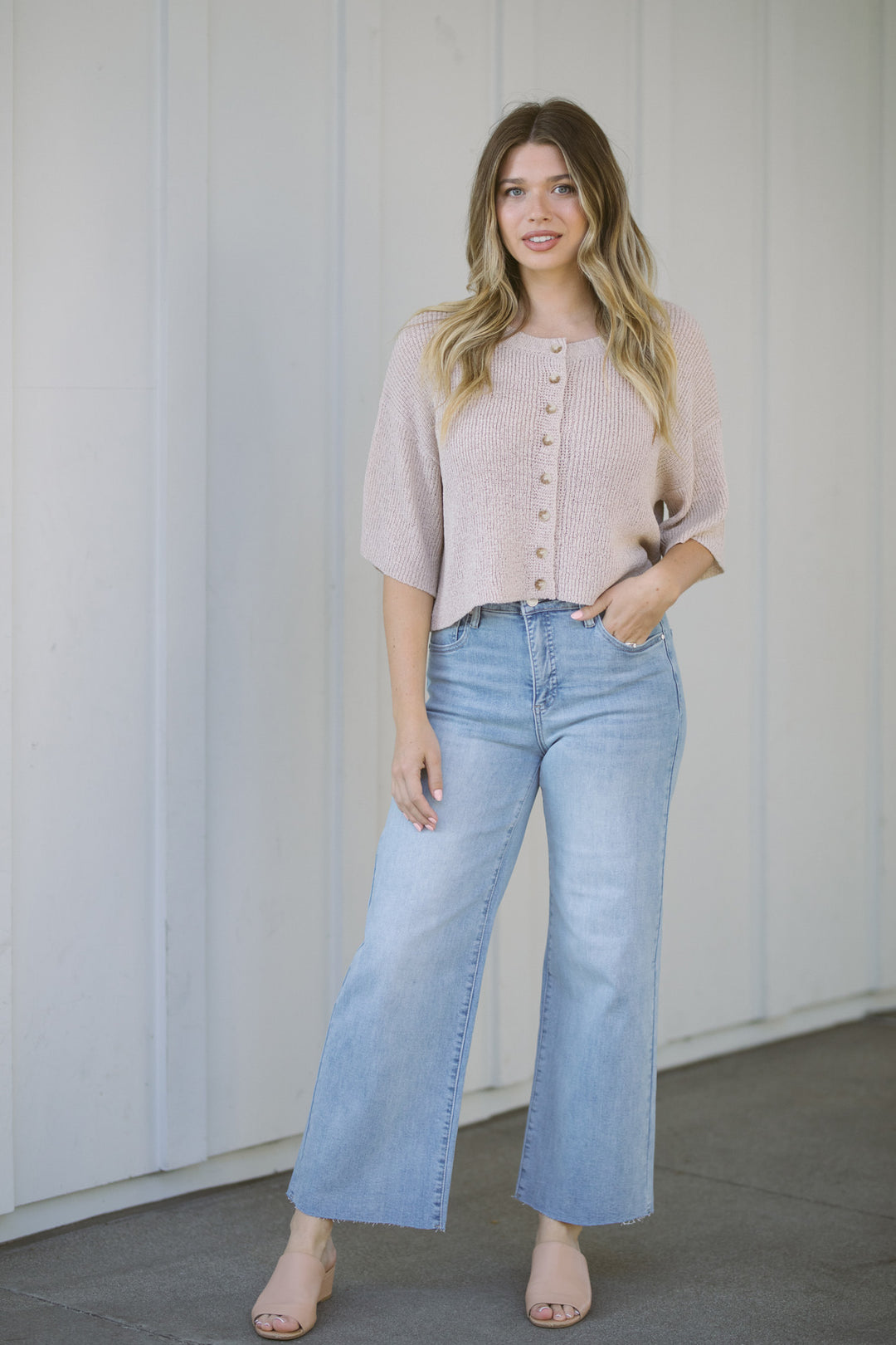 A woman wearing a beige, button-up short-sleeve cardigan and high-rise light blue wide-leg jeans, standing against a white backdrop.
