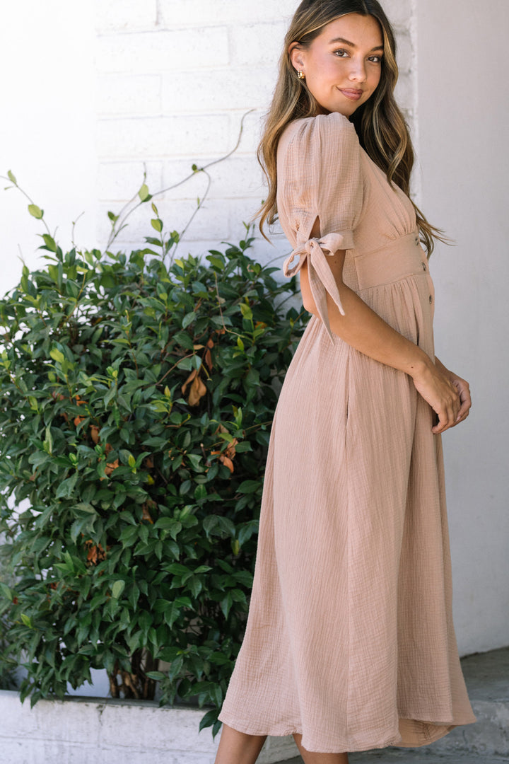 A pink, long, gauzy dress with puffed sleeves, a tie waist, and a flowing, A-line silhouette, modeled by a woman with long brown hair.