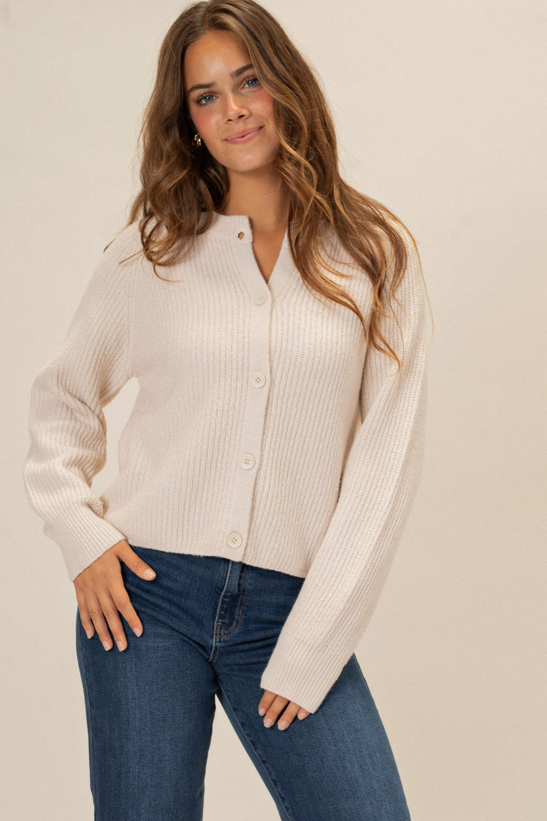 A young woman models a beige, ribbed-knit buttoned cardigan with long sleeves. She stands with her arms at her sides against a neutral background.