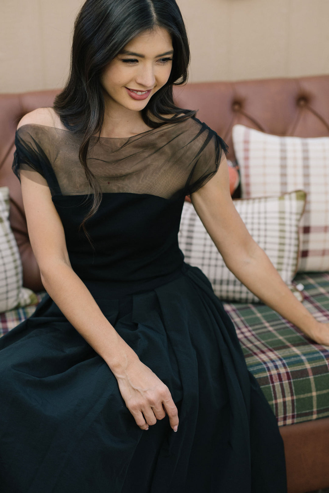 A smiling woman wearing a black mesh knit top with a draped neckline, posing on a plaid couch.