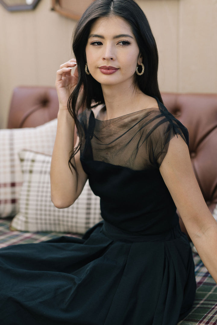 A young, attractive woman with long, dark hair wearing a black sleeveless top with a sheer, draped mesh overlay, posing confidently against a neutral background.