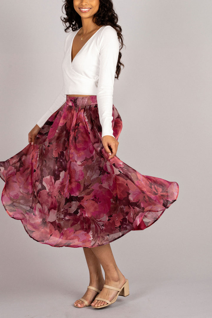 A white crop top with long sleeves is paired with a vibrant floral midi skirt in shades of pink, red, and purple. The full, flowing skirt creates a beautiful, feminine silhouette.
