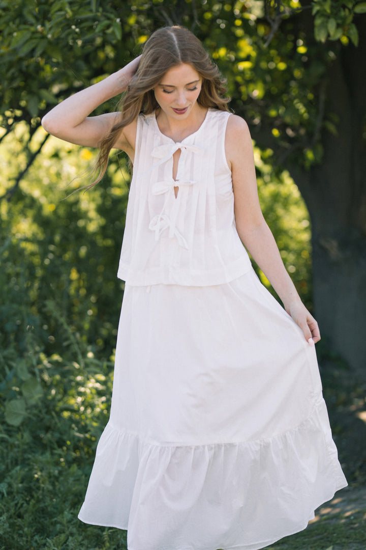 A young woman with long, wavy hair stands in a verdant outdoor setting, wearing a flowing white sleeveless dress with a tie-front detail and a tiered, midi-length skirt.