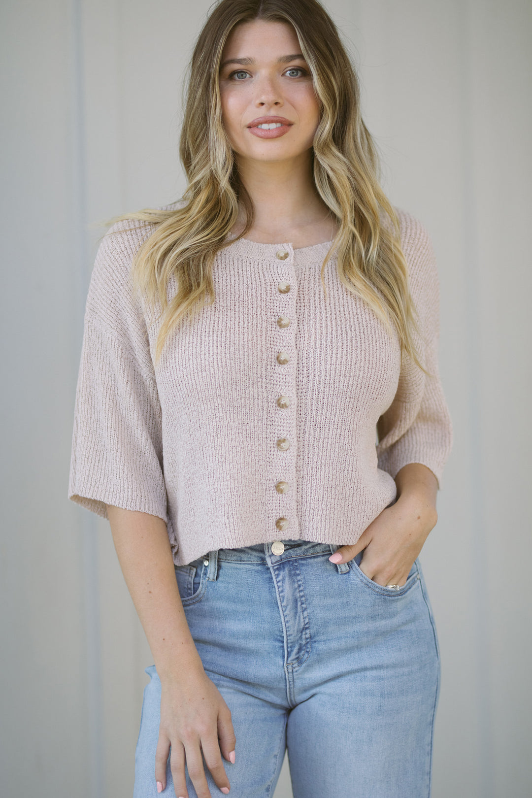 Young woman wearing a beige short-sleeve knit cardigan with wooden buttons, posing against a light background.
