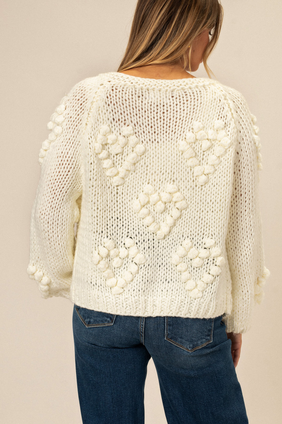 A chunky, cream-colored knit cardigan with an intricate heart-shaped pattern across the back, featuring long sleeves and a relaxed fit.